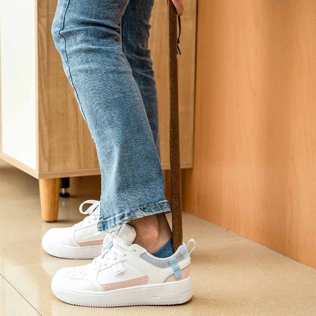 Person i jeans tar på sig vita sneakers stående med hjälp av det långa skohornet i trä